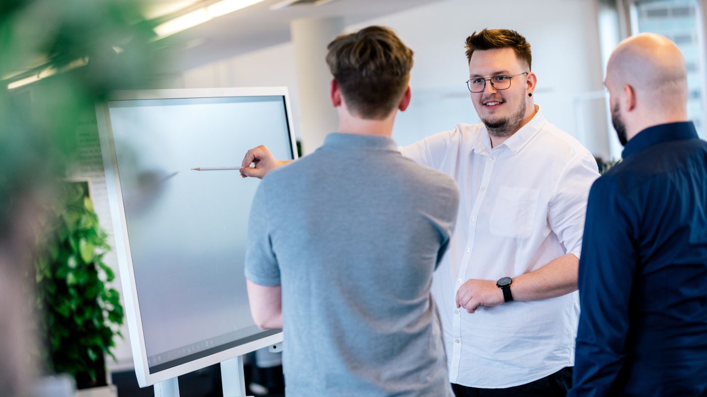 6 Schritte zu einer WordPress-Webseite Drei Personen im Gespräch. Einer der drei Präsentiert an einem Bildschirm