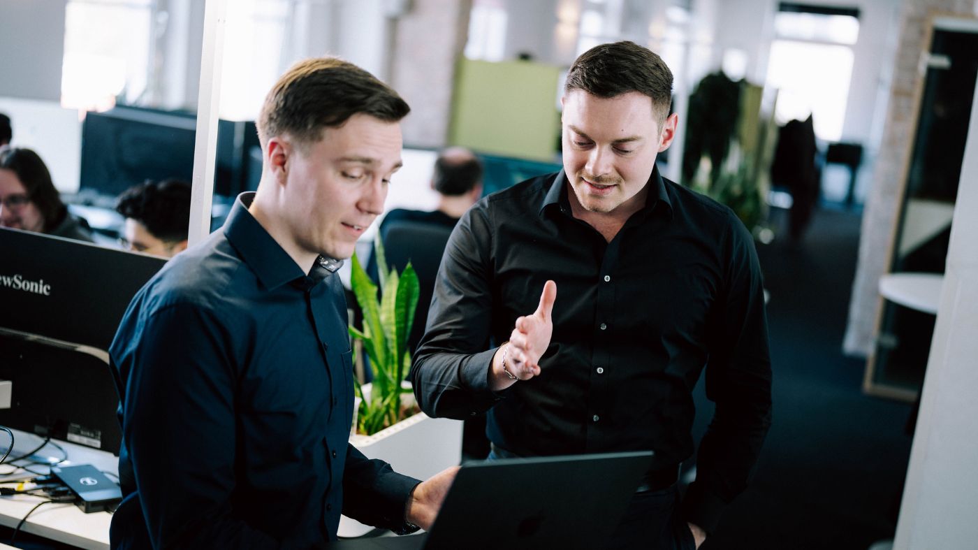 Top-5 WordPress Plugins Zwei Mitarbeiter Unterhalten sich. Einer der beiden hält einen Laptop.