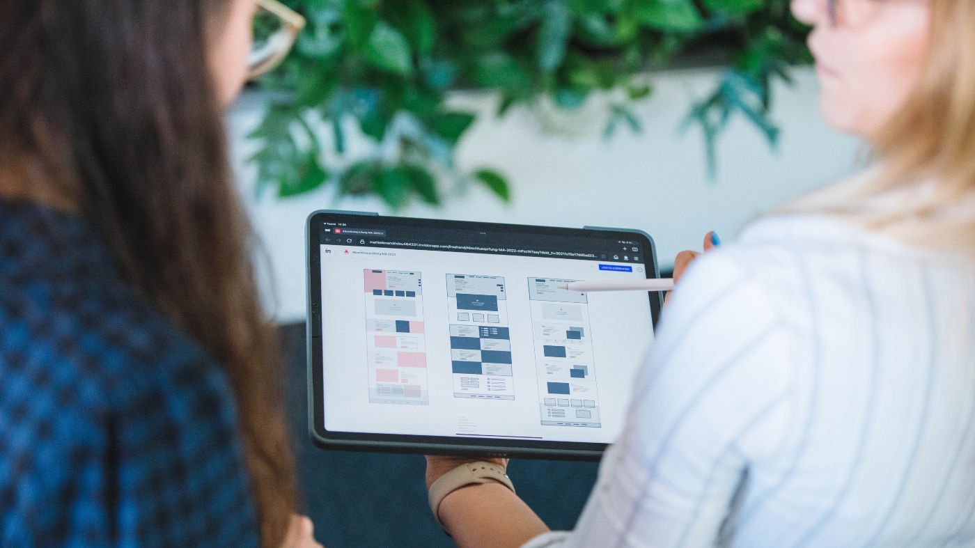 WordPress Plugins Zwei Frauen unterhalten sich. Eine der beiden hält ein Tablet und präsentiert ein Wireframe